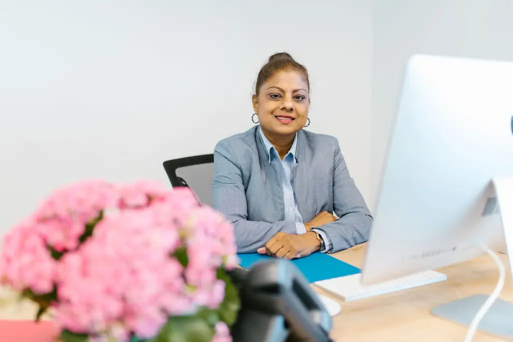 A woman in a blue suit smiling at her office desk, with flowers in the foreground