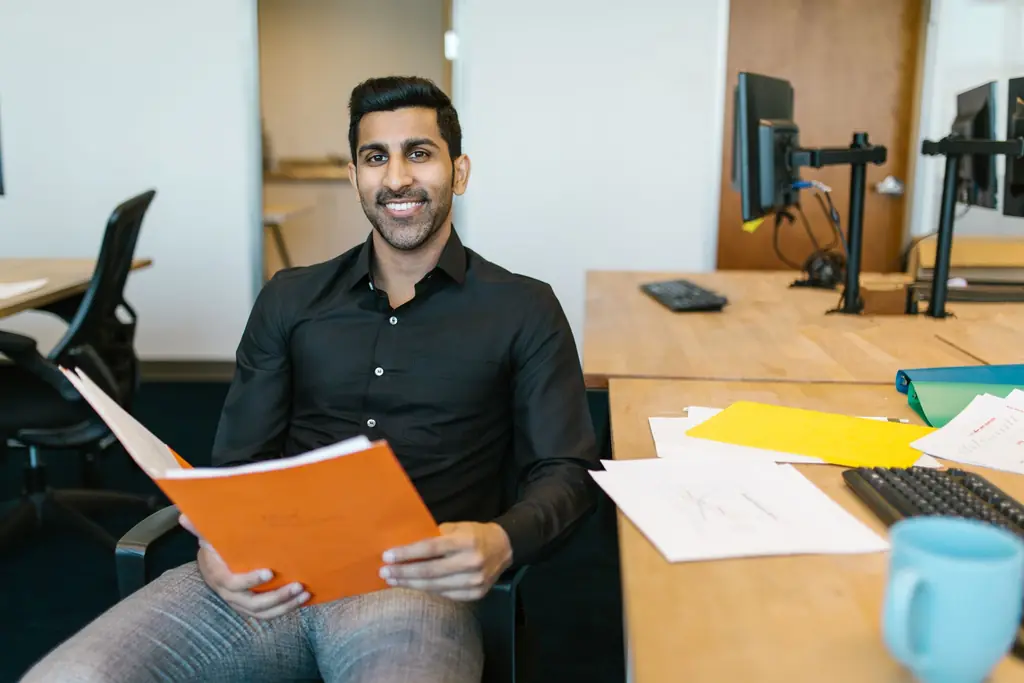 A man at his office desk holding an orange folder, smiling at the camera
